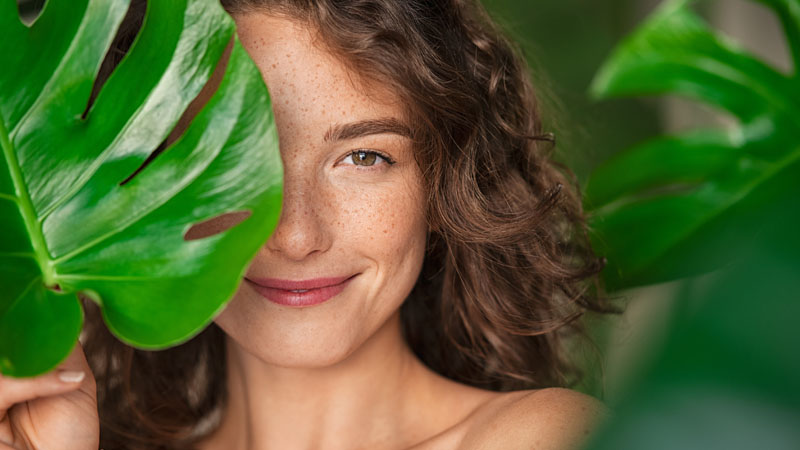 Grande plano de rosto de mulher sorridente parcialmente escondido atrás de uma planta com folhas grandes, aludindo a cosméticos naturais